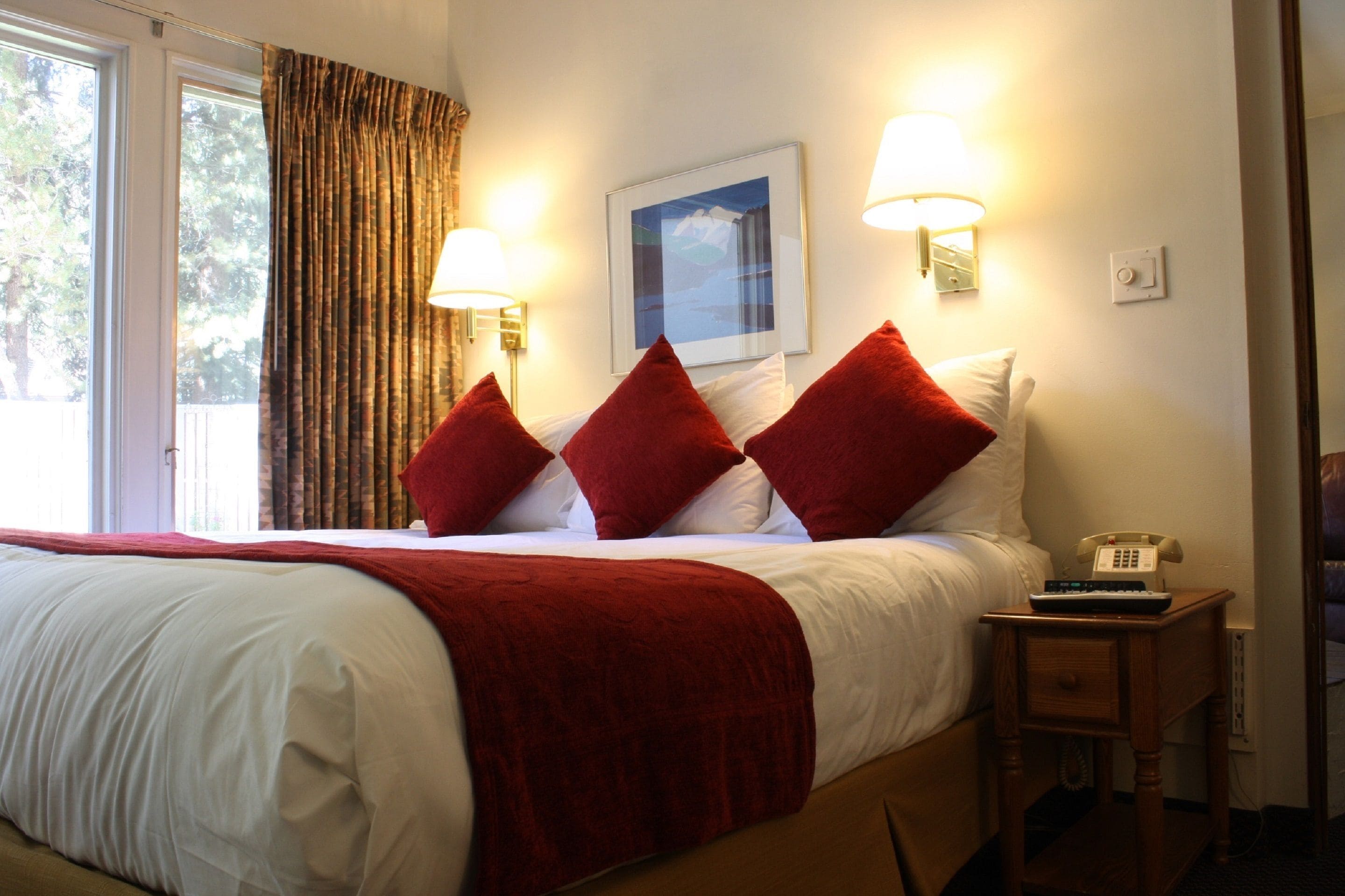 Habitación de hotel con cama, almohadas rojas, mesita de noche con teléfono y dos lámparas fijadas a la pared.