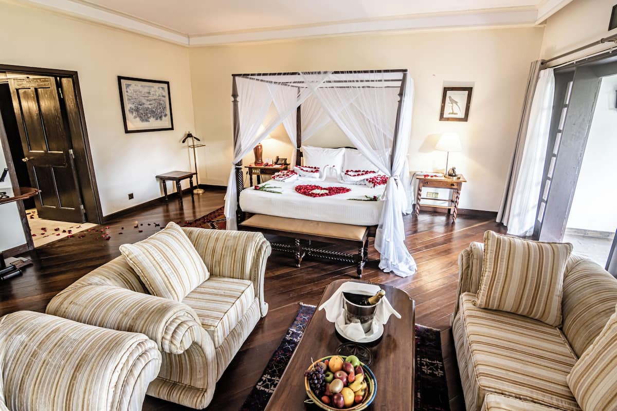 A hotel room with a four-poster bed, two striped sofas, a coffee table with fruit, and a bedside table with a lamp.