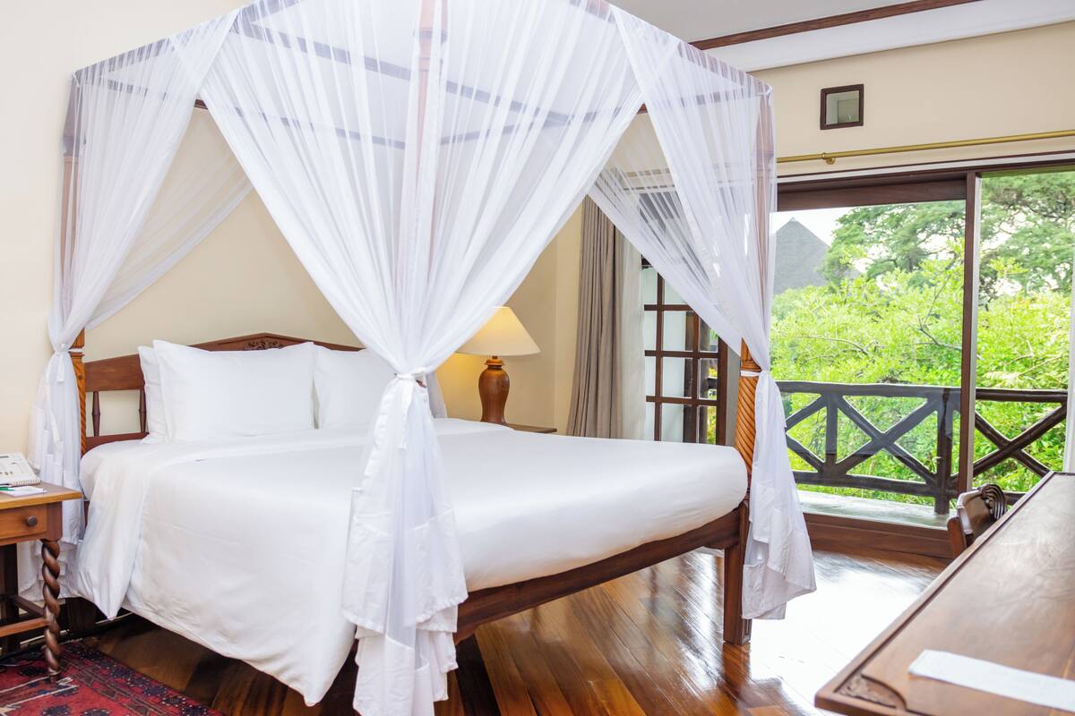 A bed with a canopy, a bedside table, a lamp, and a view of greenery through a window.