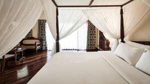 Una cama con dosel y estructura de madera, con ropa de cama blanca. Hay una silla de madera con respaldo trenzado y una mesita auxiliar a su lado. El piso es de madera oscura y las cortinas de las ventanas tienen un diseño estampado.