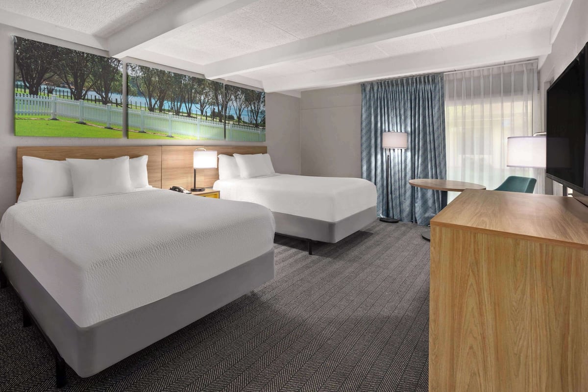 A hotel room with two beds, a desk, a TV, and a large window with curtains.