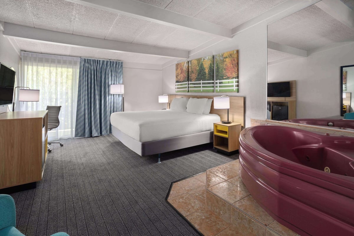 A hotel room with a bed, a desk, a chair, a TV, a large window with curtains, a painting, a jacuzzi, and a mirror.