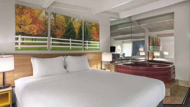 A large bed with white linens, a wooden headboard, and two bedside lamps. There is a red hot tub in the background, and a large mirror reflecting the room. - Days Inn by Wyndham Ann Arbor (Ann Arbor)