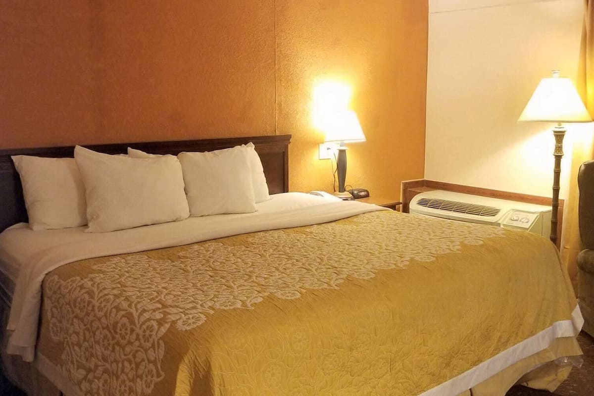 A hotel room with a bed, two bedside lamps, a chair, and an air conditioning unit.