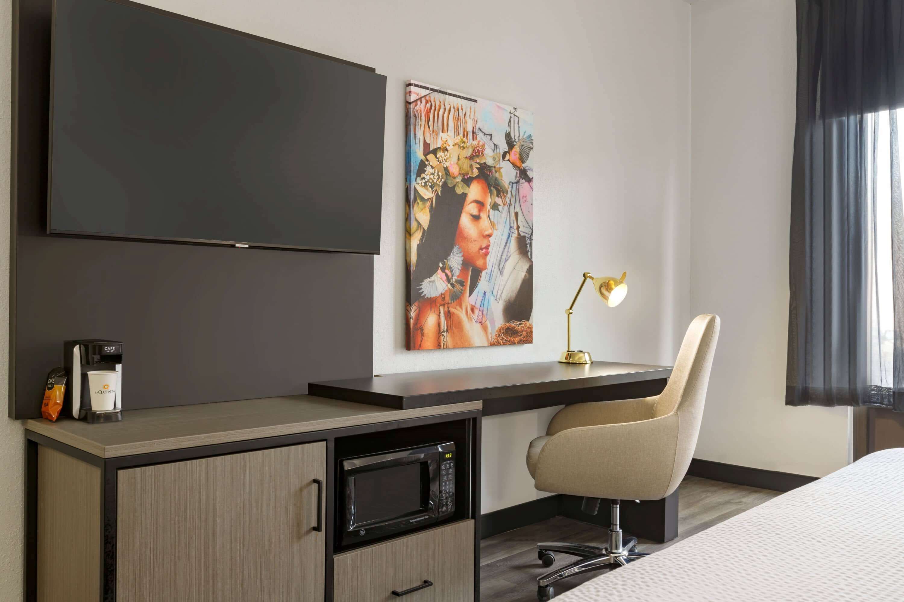 Habitación de hotel con televisión de pantalla plana, un cuadro, un escritorio con lámpara, una silla, microondas y una cama.