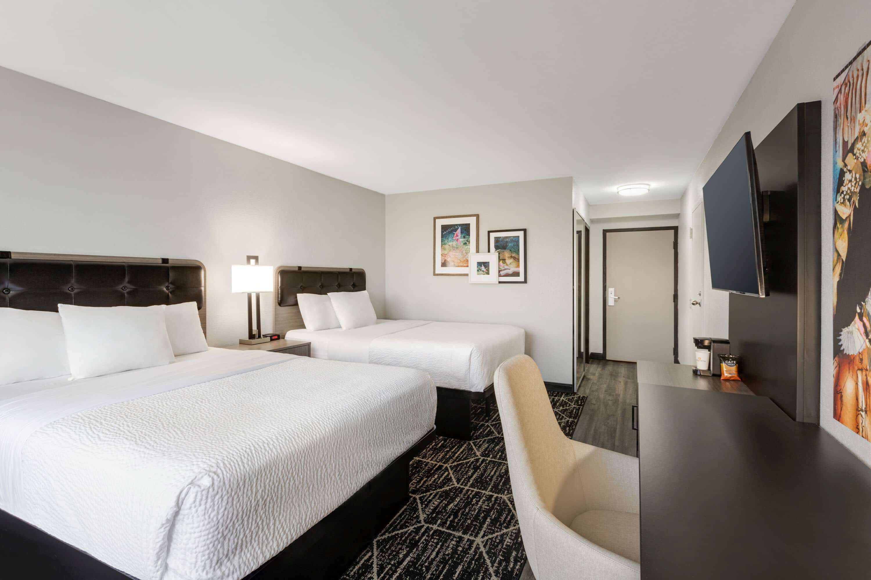Habitación de hotel con dos camas, un escritorio, una silla, un televisor y obras de arte en la pared.