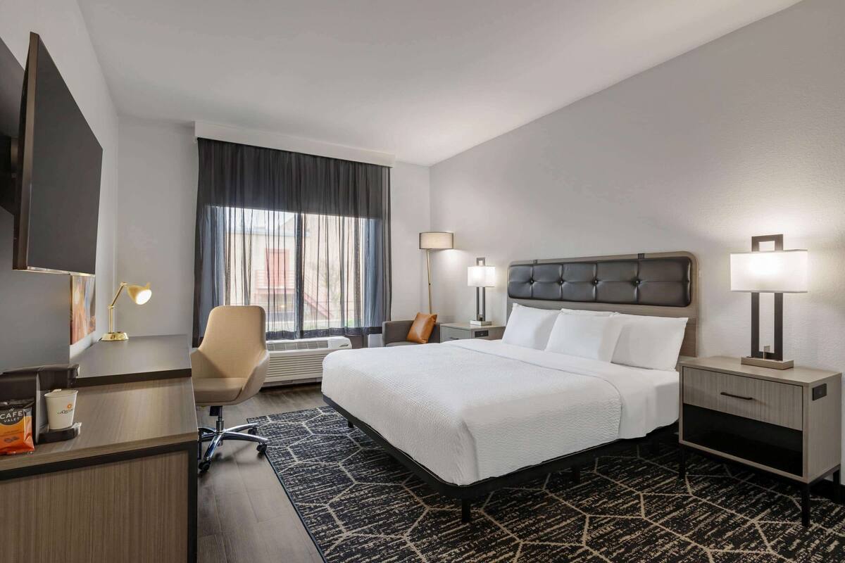 A hotel room with a large bed, a desk with a lamp, a chair, a nightstand with a lamp, and a window with curtains.