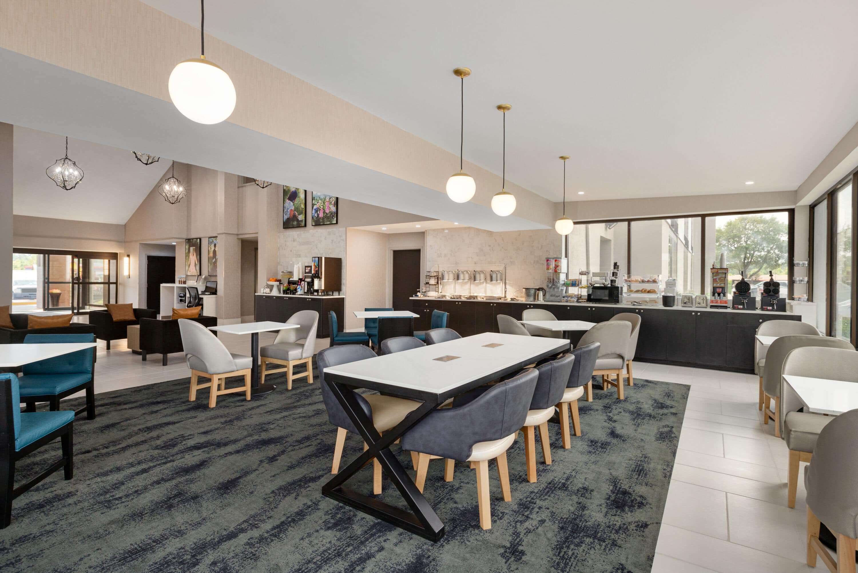 Una hall d'albergo moderna con un'area ristorazione centrale, un bar e una cucina visibili attraverso una parete di vetro.