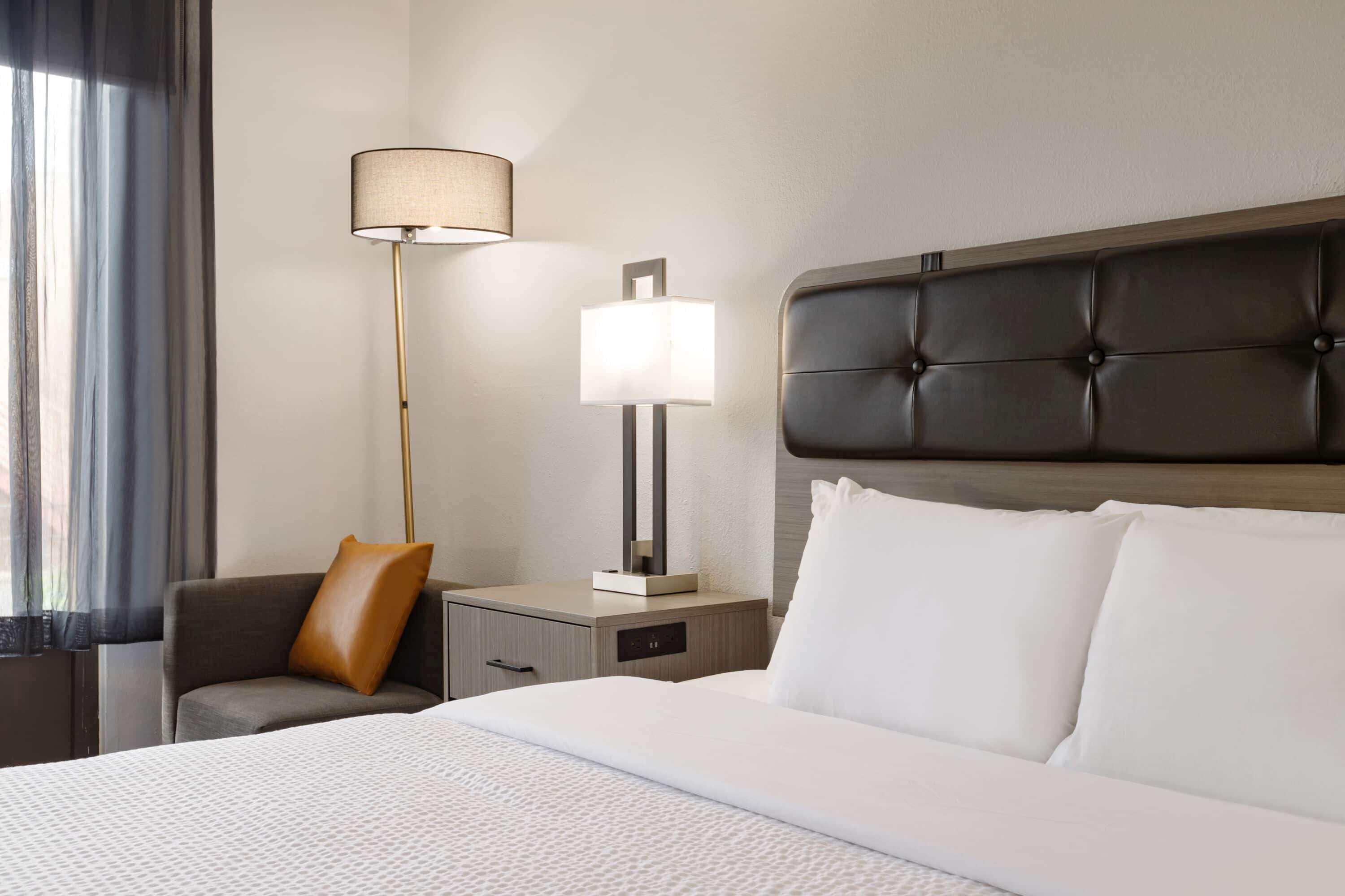 Una habitación de hotel con una cama grande, una mesita de noche, una lámpara, una silla y una ventana con cortinas.