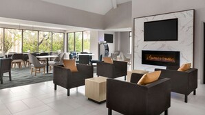 A spacious living area with dark sofas, a fireplace, a flat-screen TV, and large windows offering a view of greenery. - La Quinta Inn & Suites by Wyndham Dothan (Dothan)