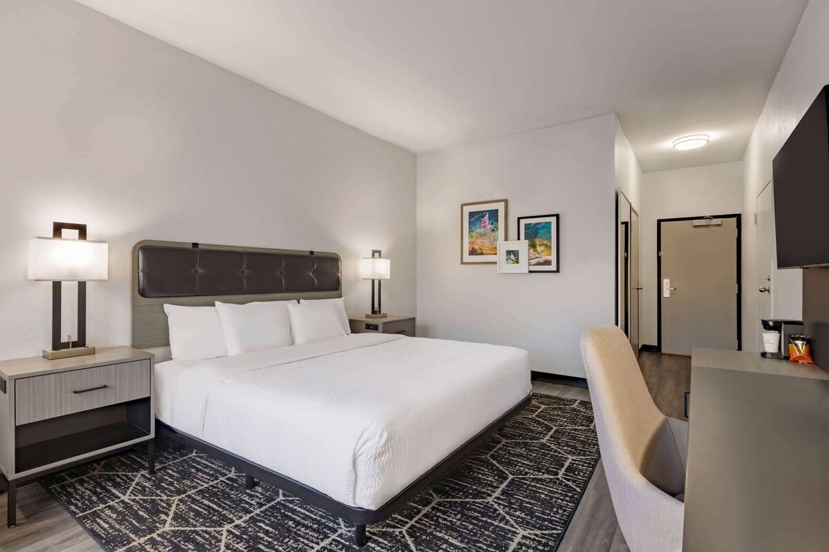 A hotel room with a large bed, bedside tables with lamps, a desk, and a chair. There are framed pictures on the wall and a door in the background.