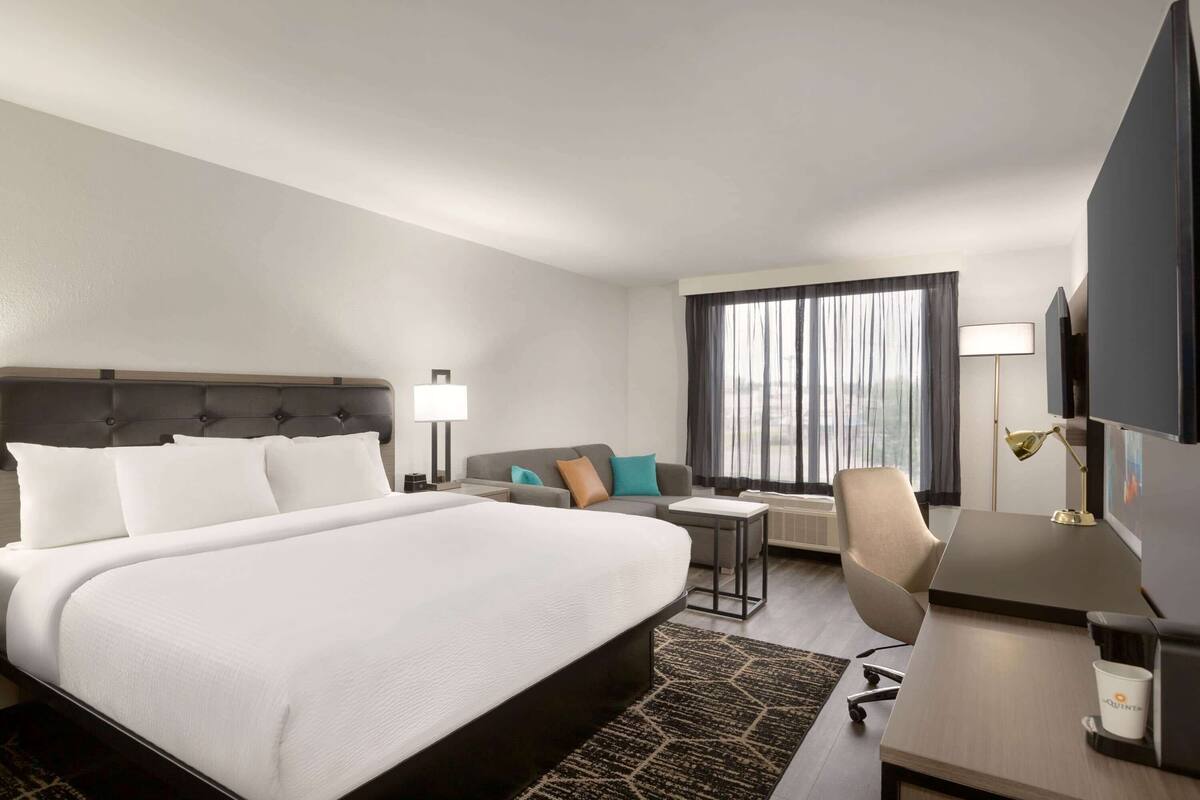 A hotel room with a large bed, a desk, a chair, a sofa, a TV, and a window with curtains.