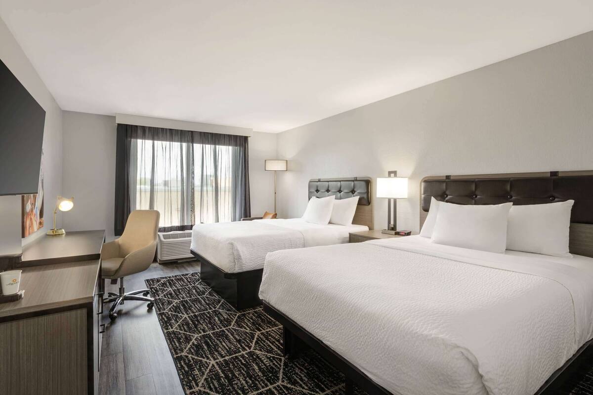 A hotel room with two beds, a desk, a chair, a lamp, and a large window with curtains.