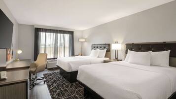 A hotel room with two beds, a desk, a chair, a lamp, and a large window with curtains.