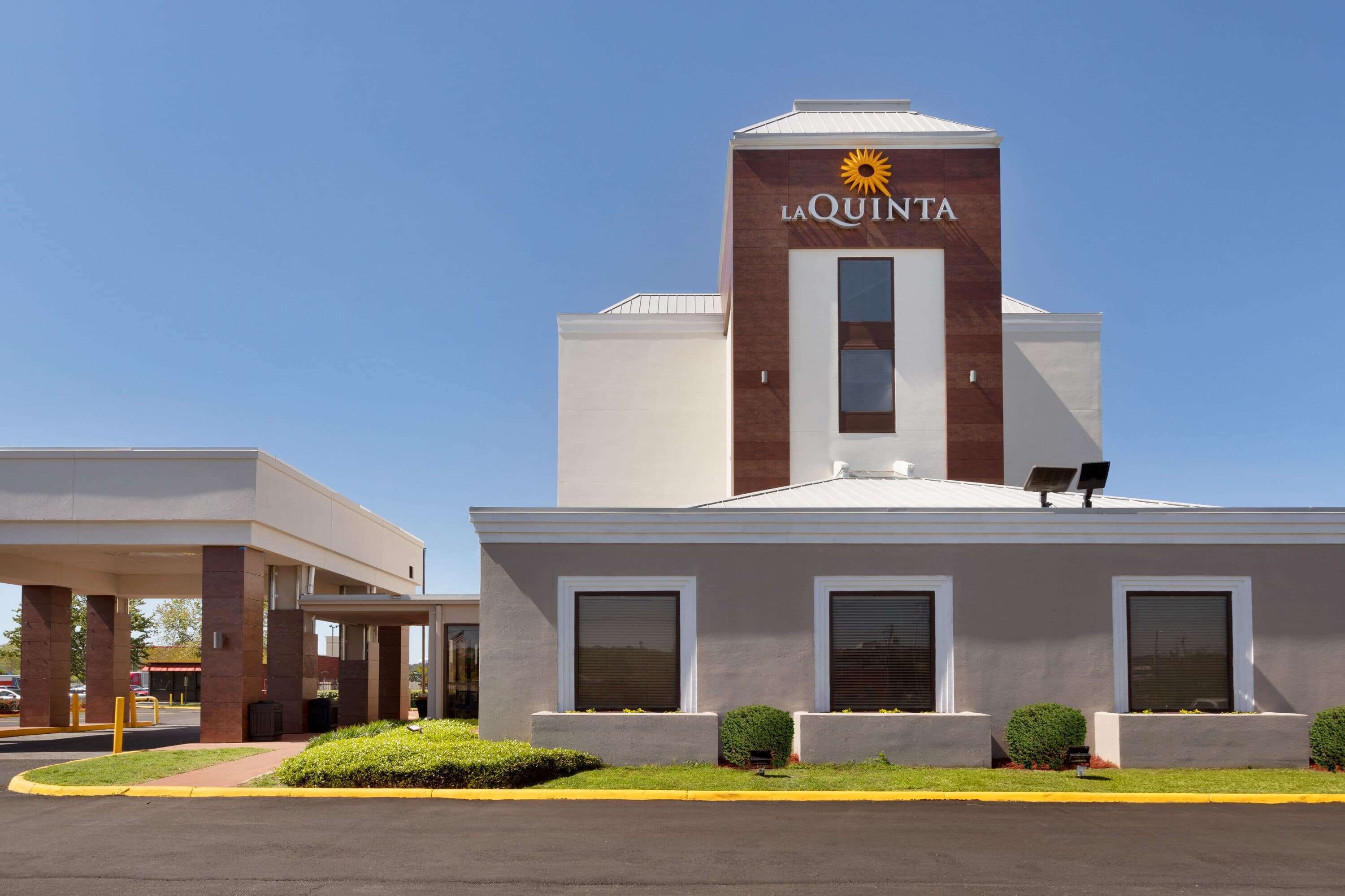 A La Quinta hotel with a modern design, featuring a prominent sun logo on the facade, a covered parking area, and a well-maintained lawn.