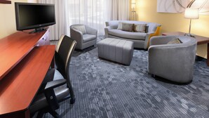 Quarto de hotel com televisão de tela plana, escrivaninha de madeira, sofá cinza, poltrona cinza, banquinho cinza e luminária de pé.