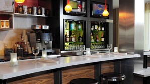 Uma área de bar com bancada branca, dois banquinhos, uma máquina de café e prateleiras repletas de garrafas e copos.