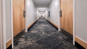 Un long couloir étroit, typique d’un hôtel, avec des revêtements de sol en moquette, des portes en bois et des murs de couleur claire.