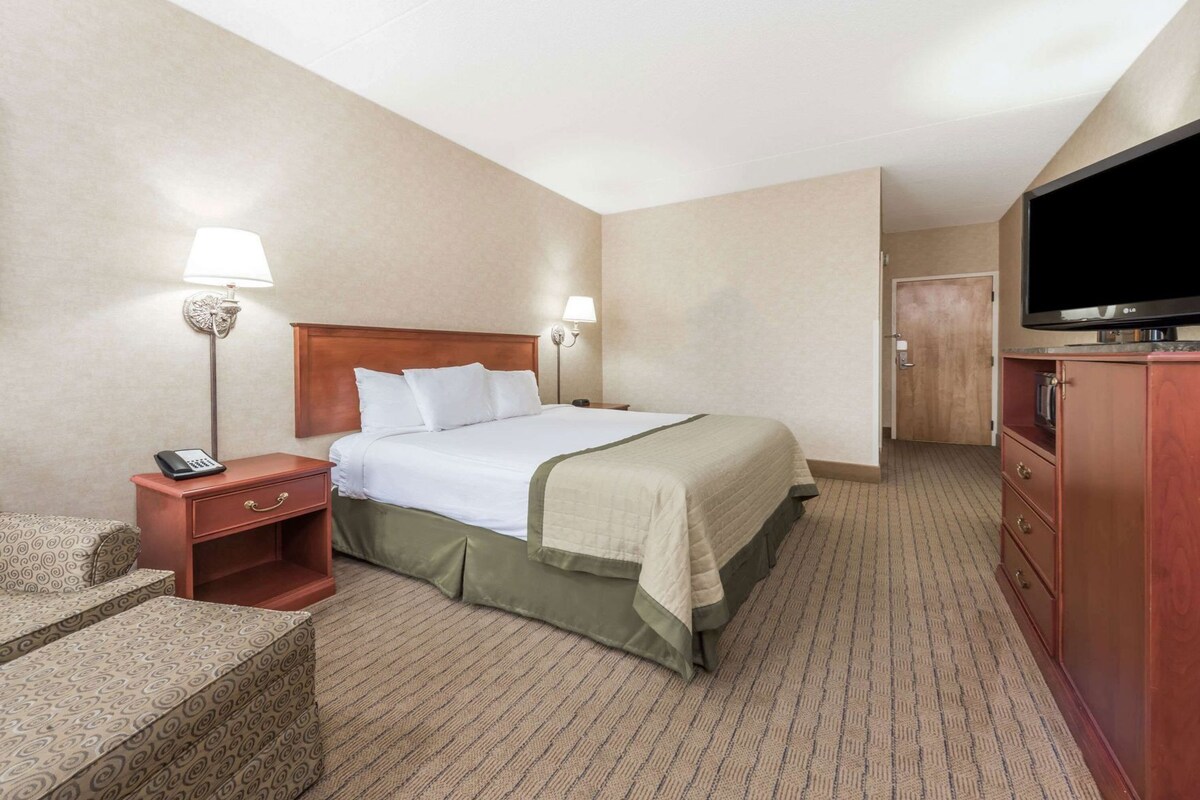 A hotel room with a bed, bedside lamps, a desk with a telephone, a television on a stand, and a door leading to another room.
