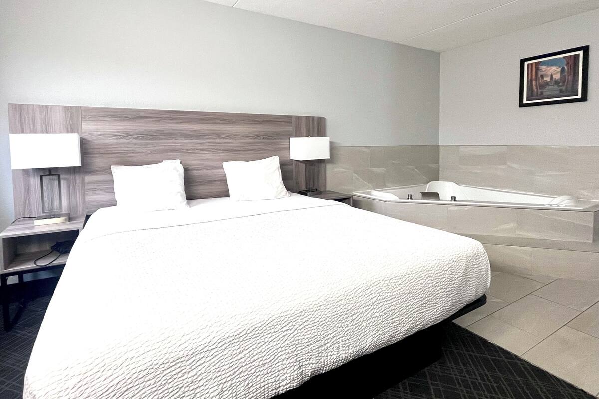 Een hotelkamer met een groot bed, twee nachtkastjes met lampen, een ingebouwd bad en een ingelijst kunstwerk aan de muur.