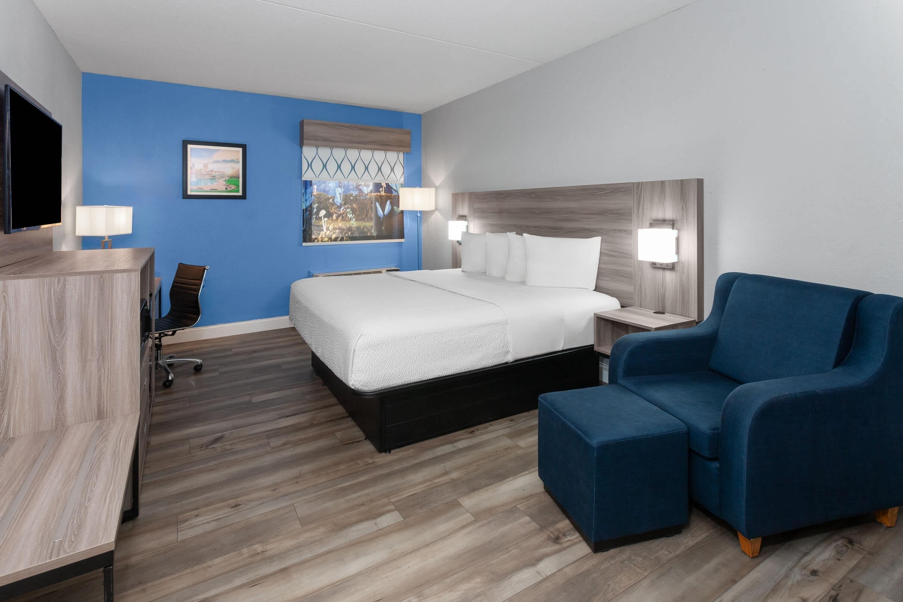Une chambre d’hôtel avec un grand lit, un bureau avec une chaise, un fauteuil bleu et un téléviseur posé sur un bureau en bois.