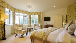 A bedroom with a large bed, two armchairs, a small table, a dresser, a television, and a mirror. - Hare And Hounds Hotel (Tetbury)