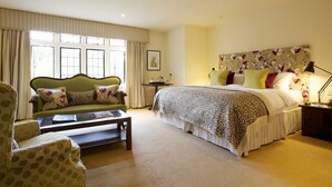 A bedroom with a bed, a green armchair, a small table, and a nightstand with a lamp. - Hare And Hounds Hotel (Tetbury)