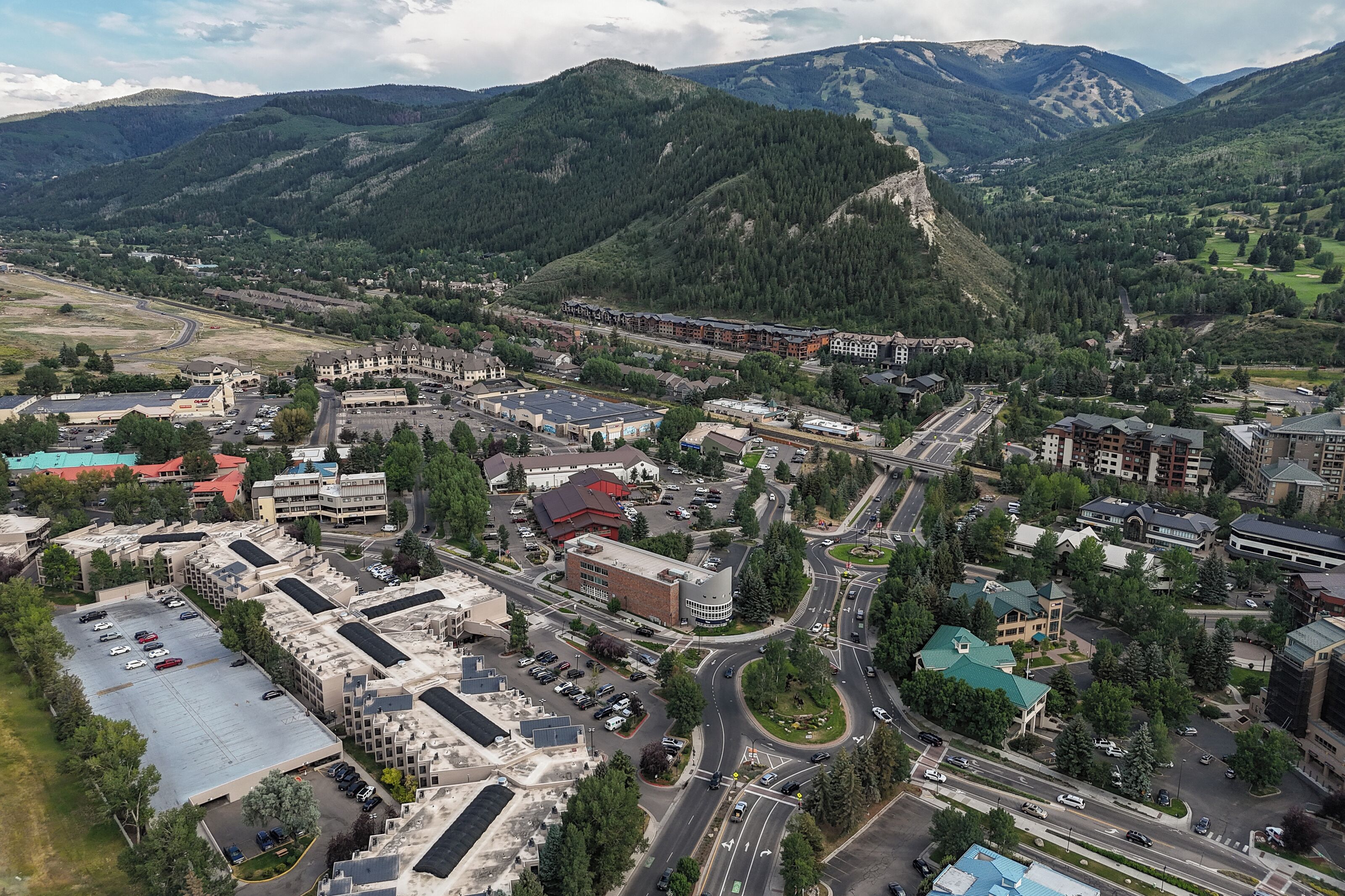 An aerial view of a town with a mix of commercial and residential buildings, roads, and a mountainous backdrop.