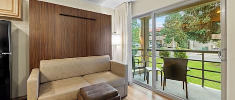 A living room with a sofa, ottoman, wooden panel wall, and a sliding glass door leading to an outdoor area with a table and chairs.