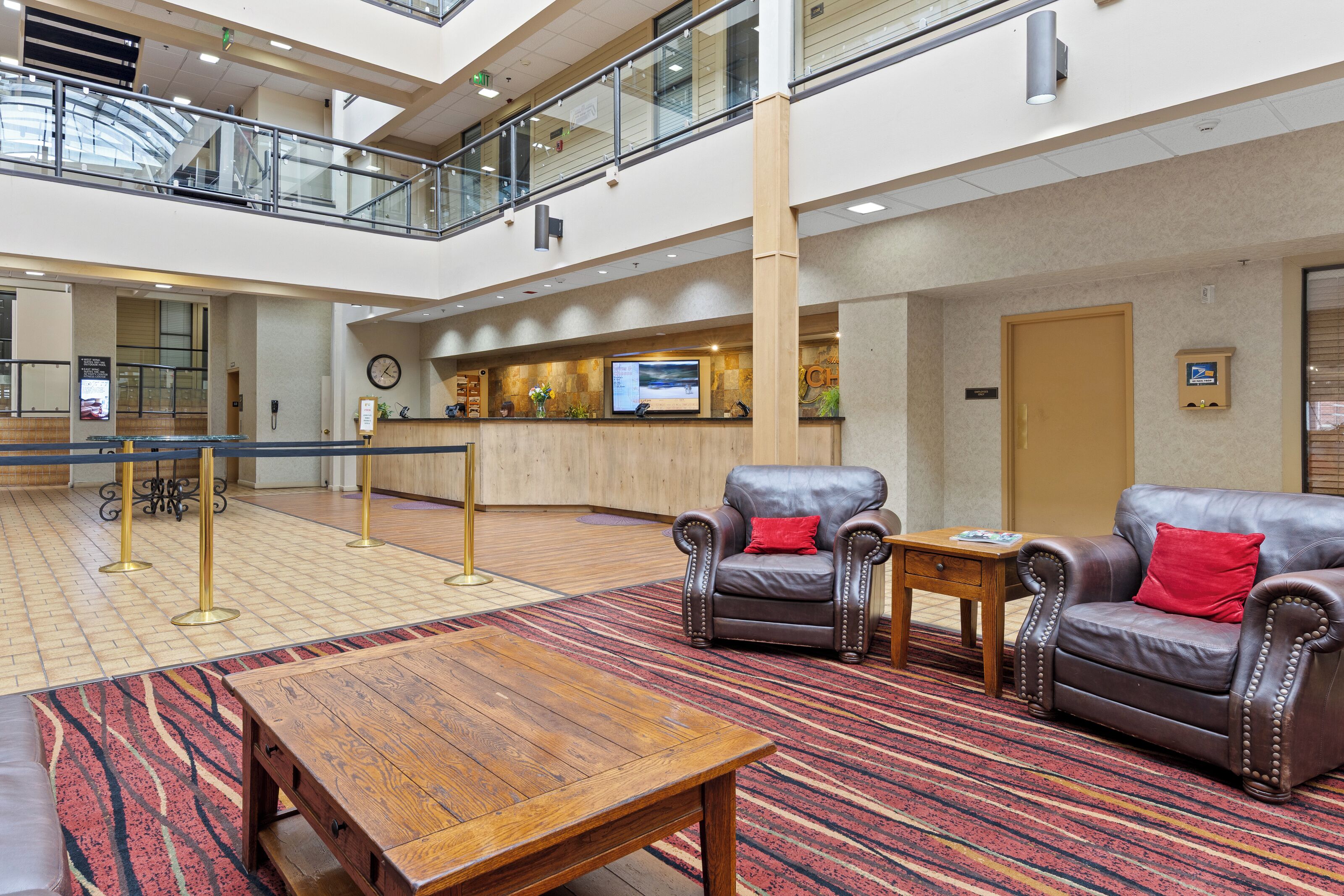 A lobby with a reception desk, two leather armchairs with red cushions, a wooden coffee table, and a patterned carpet.