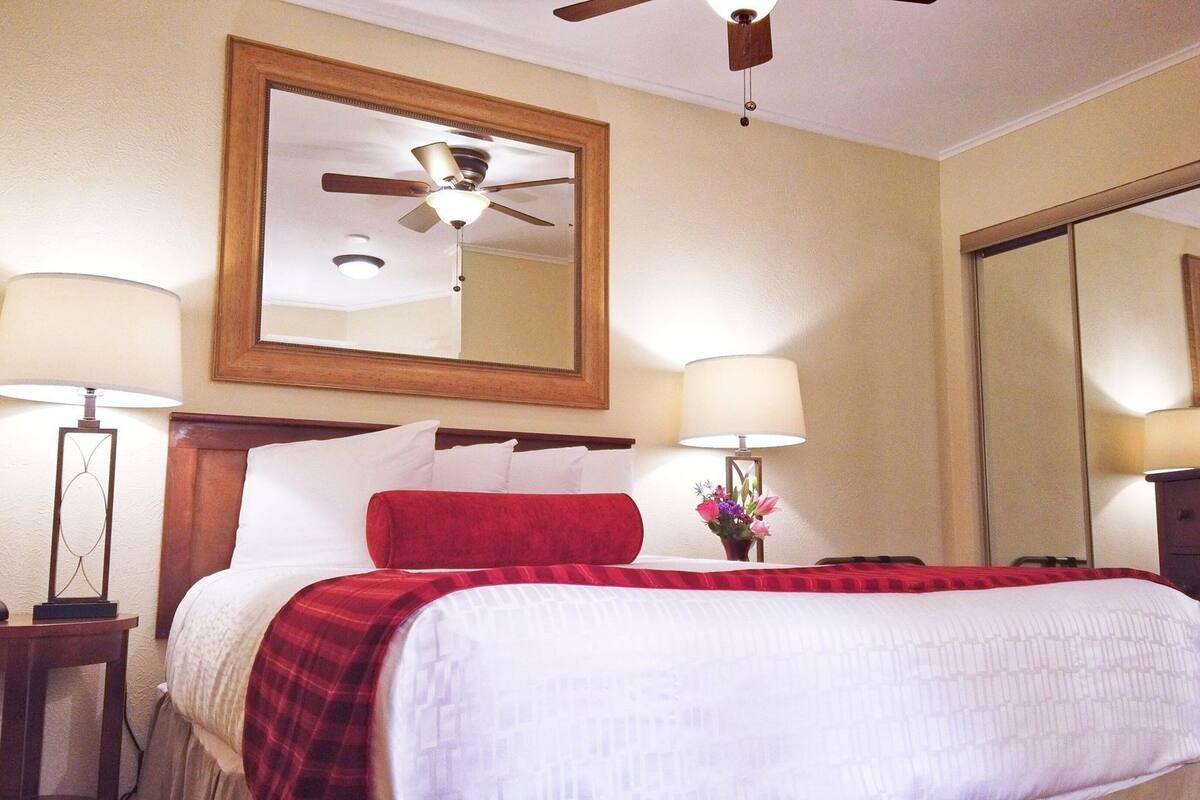A hotel room with a bed, two bedside tables, a ceiling fan, a large mirror, and a TV.