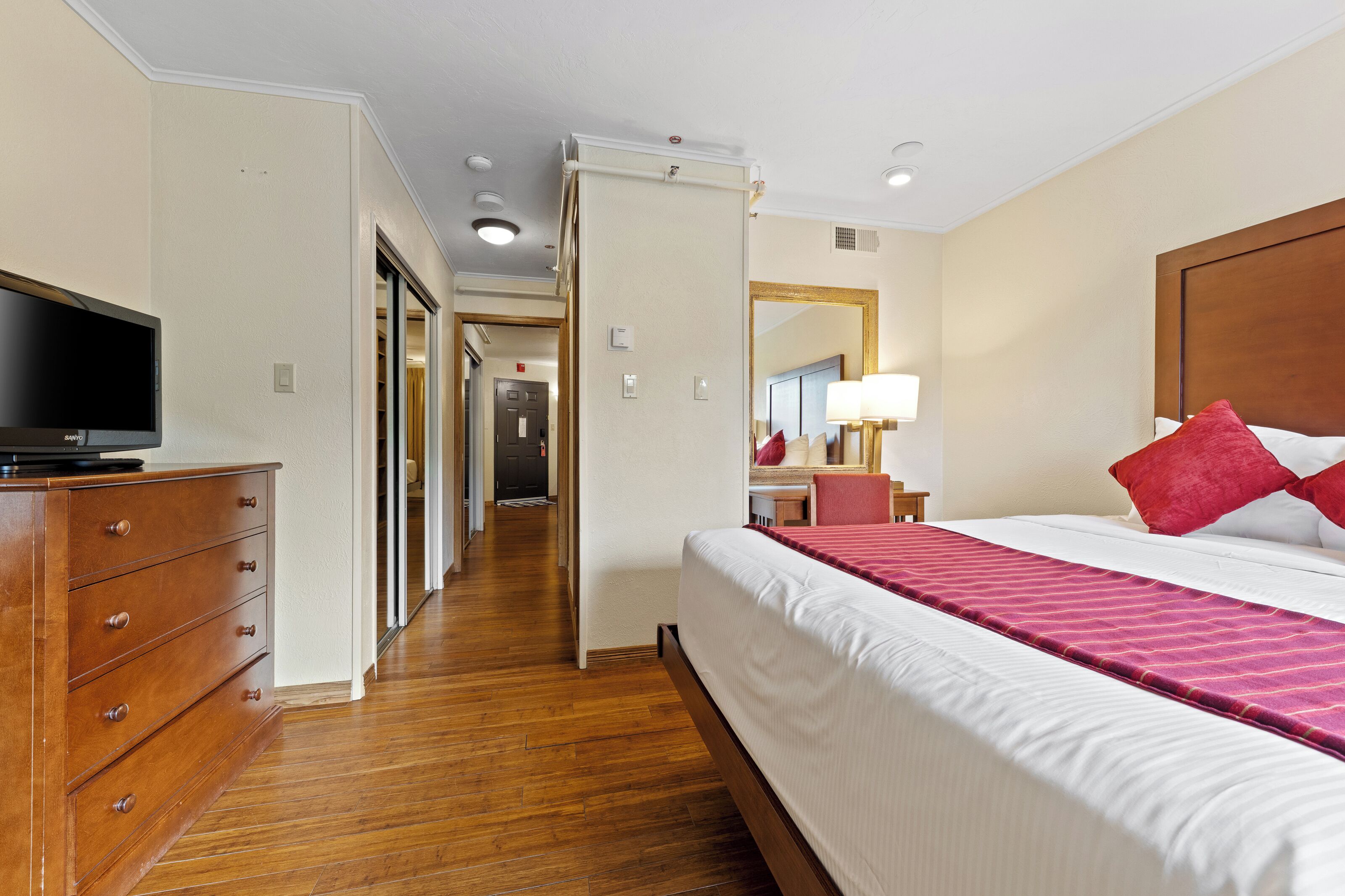 A hotel room with a wooden dresser, a flat-screen TV, a large bed with red pillows, and a mirror on the wall.