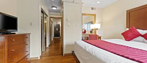 A hotel room with a wooden dresser, a flat-screen TV, a large bed with red pillows, and a mirror on the wall.