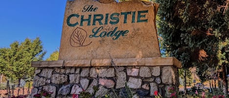 A stone sign with "The CHRISTIE Lodge" engraved on it, surrounded by lush greenery and flowering plants.