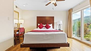 A hotel room with a large bed, a desk with a chair, a ceiling fan, and a large window with a view of the outdoors.