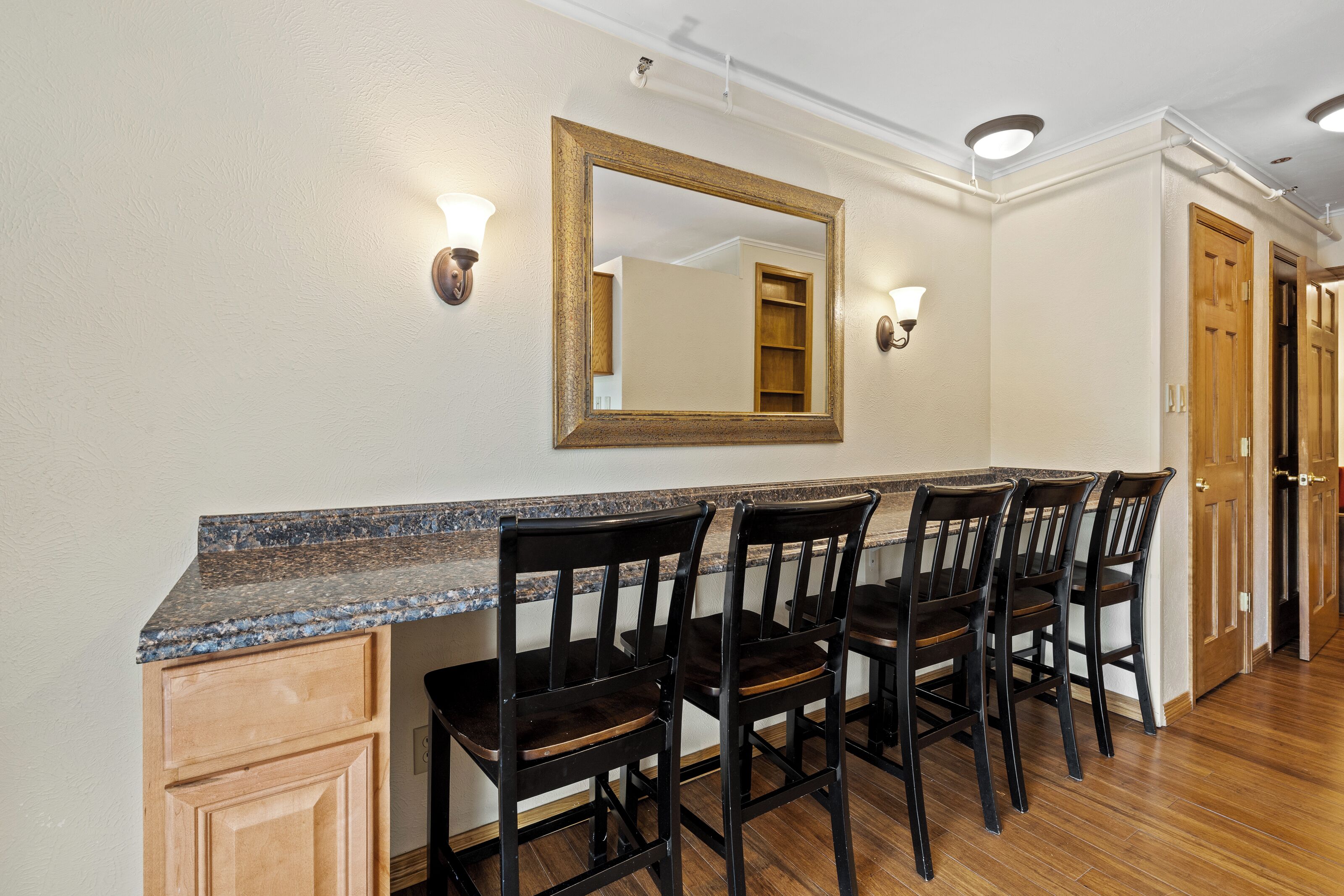 A kitchen with a granite countertop, four dark wooden chairs, and a large mirror above the counter.
