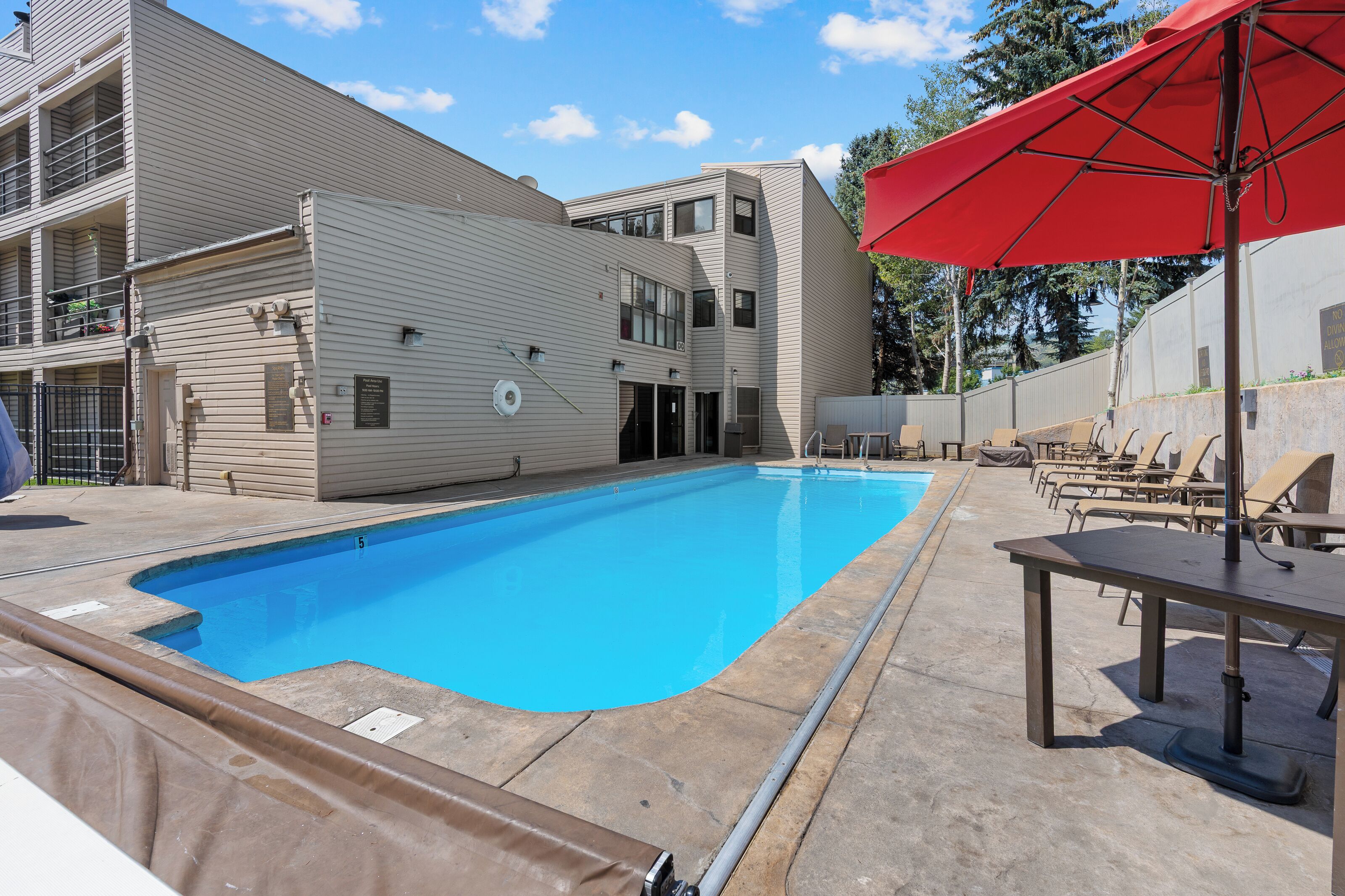 A swimming pool with a red umbrella, lounge chairs, and a building with multiple windows and balconies.