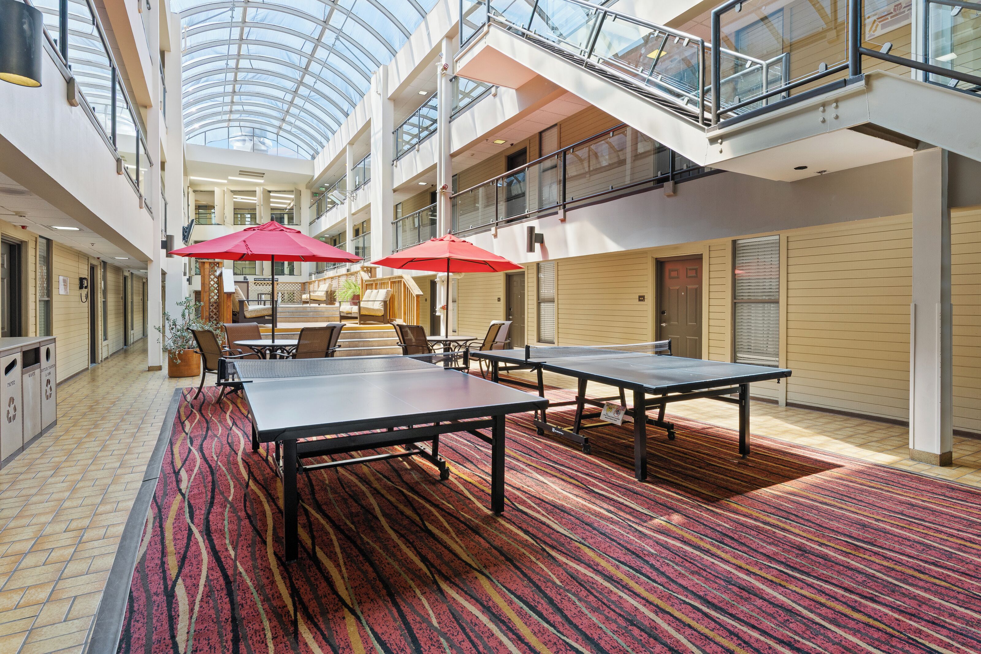 A spacious indoor area with ping pong tables, red umbrellas, and a high ceiling with a glass skylight.