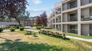 A modern apartment building with multiple balconies, a grassy courtyard with picnic tables, and a parking area.