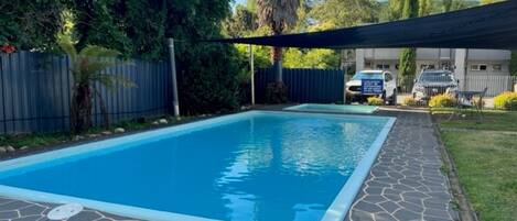 A rectangular swimming pool with a stone border, surrounded by a fenced yard with trees and a car parked in the background.