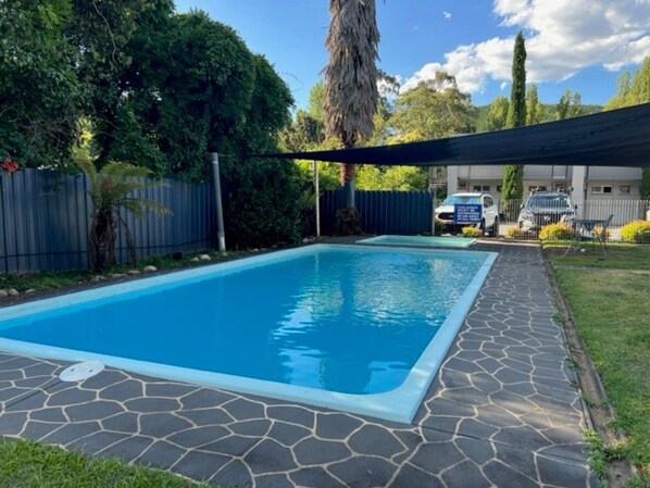 A rectangular swimming pool with a stone border, surrounded by a fenced yard with trees and a car parked in the background. - Riverbank Park Motel (Bright)