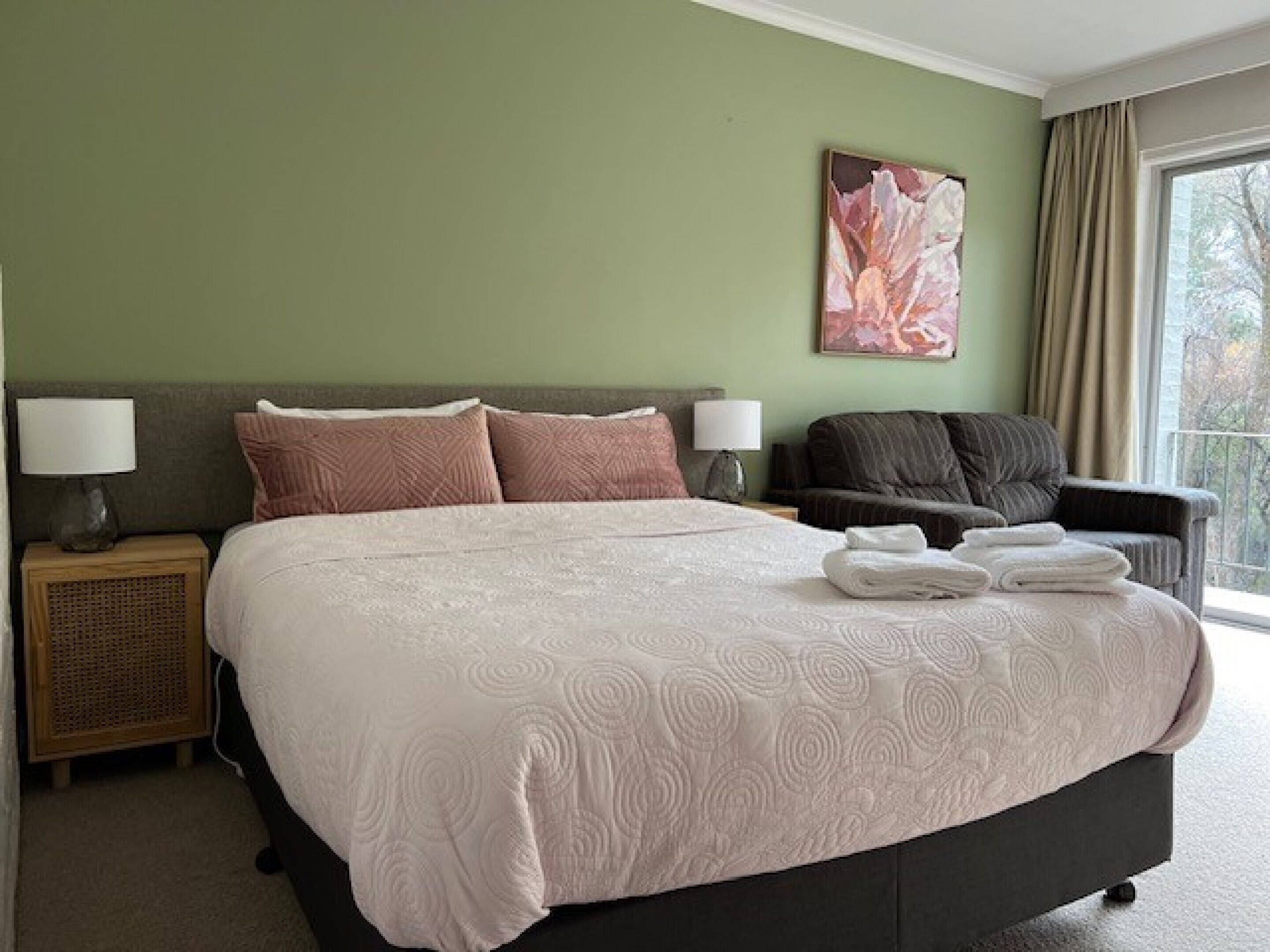 A hotel room with a large bed, two bedside tables with lamps, a dark armchair, and a large window with curtains.