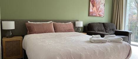 A hotel room with a large bed, two bedside tables with lamps, a dark armchair, and a large window with curtains.