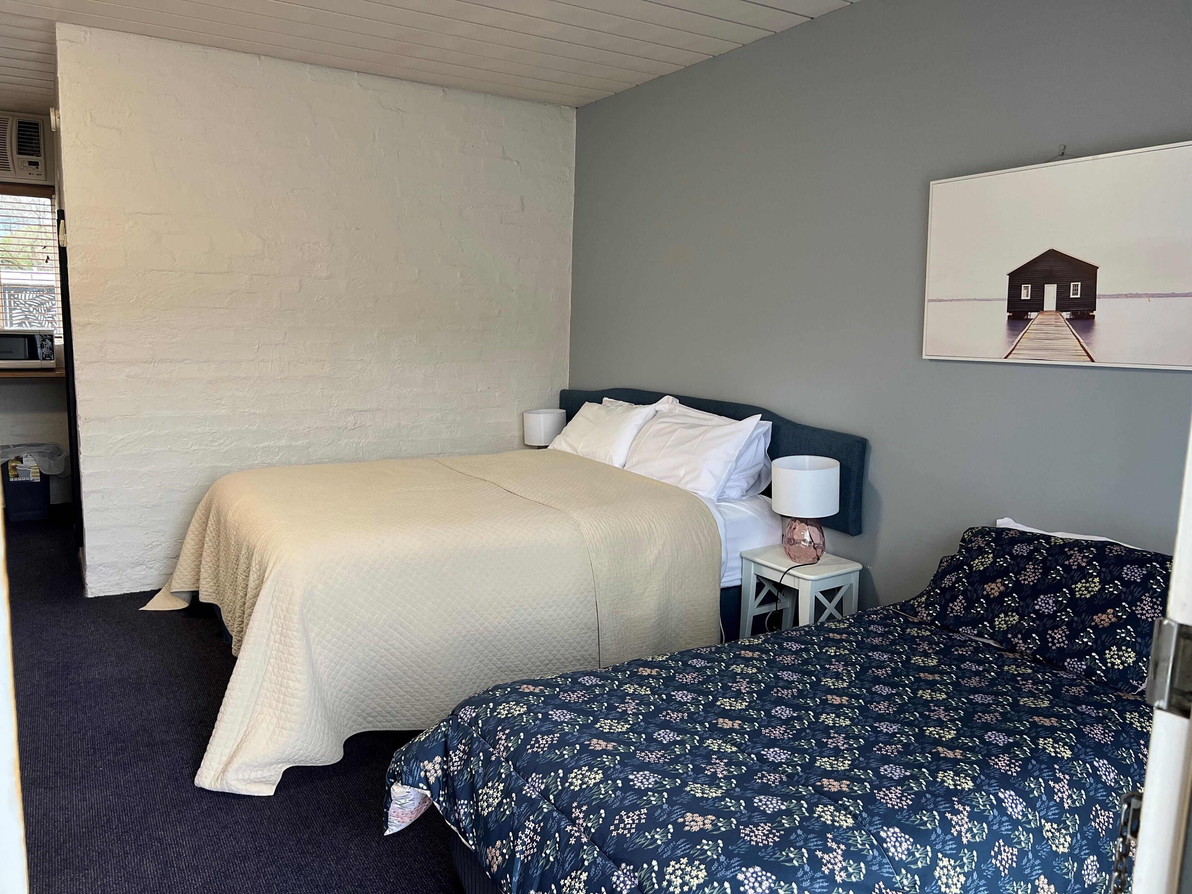 A bedroom with a bed, a bedside table with a lamp, a wall-mounted picture, and a window with blinds.