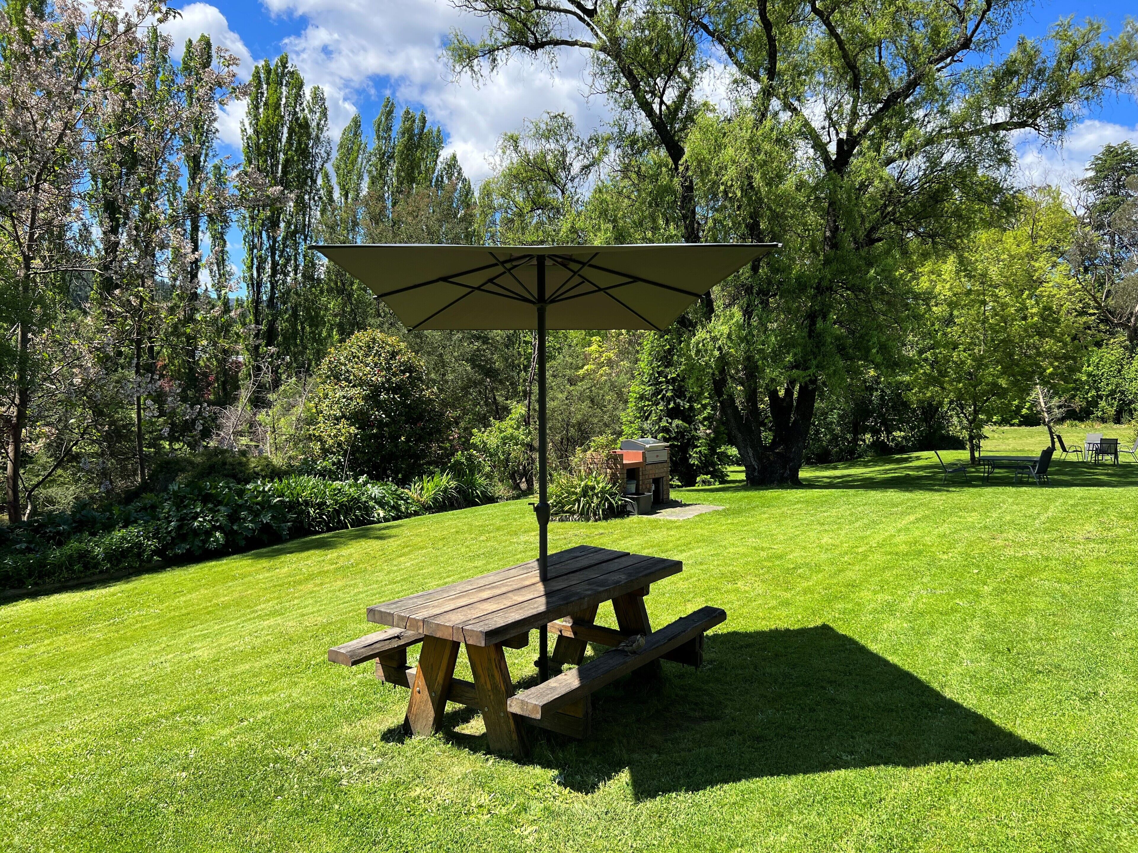 Ein Picknicktisch mit Sonnensegel in einem gepflegten Grünbereich mit Bäumen und einem Grill im Hintergrund.