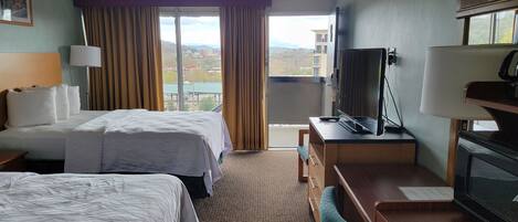 Habitación de hotel con dos camas, un escritorio con televisor y vista a la ciudad desde la ventana.