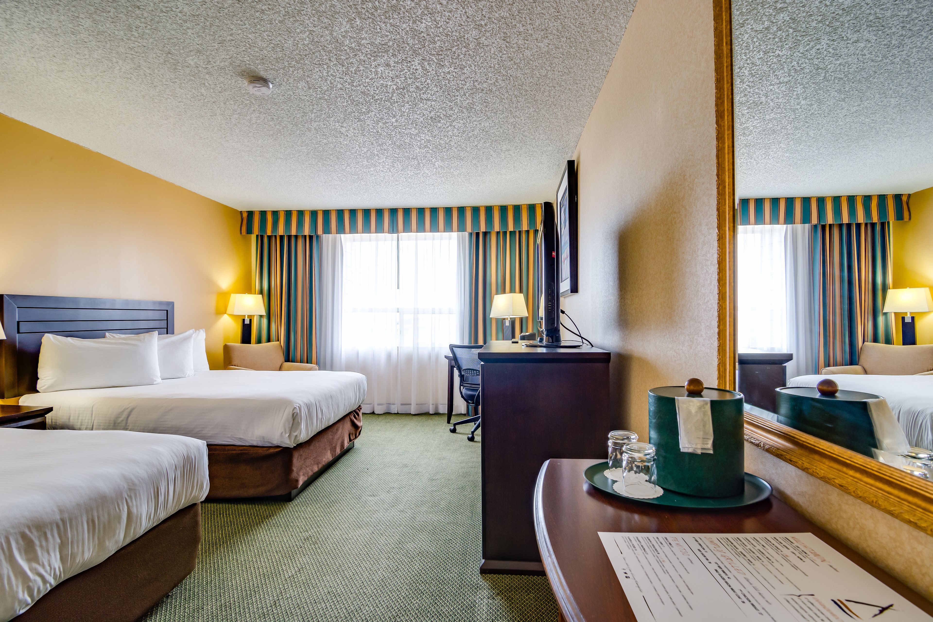 A hotel room with two beds, a desk, a mirror, and a green carpet.