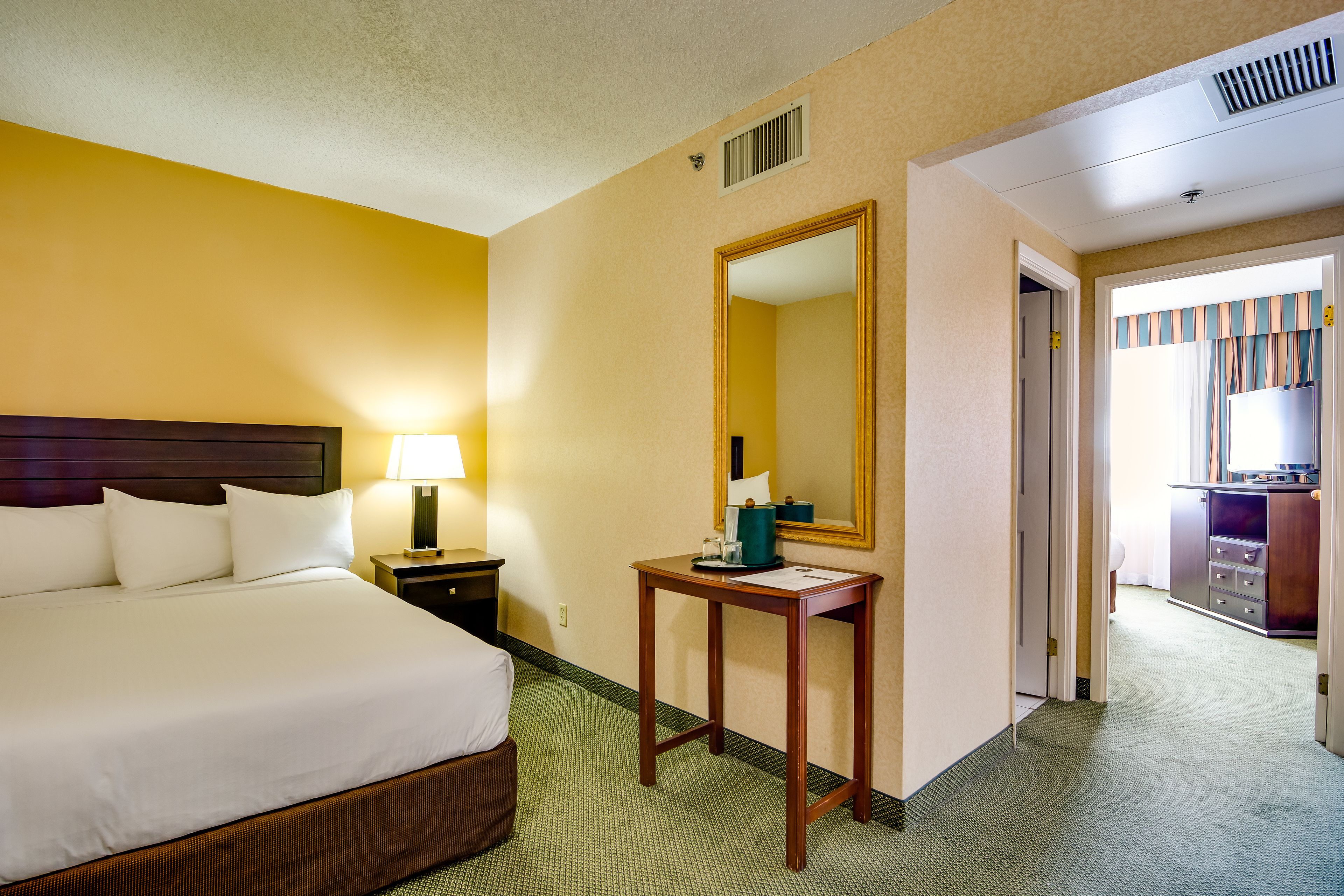 A hotel room with a large bed, a bedside table, a mirror, and a desk with a chair.