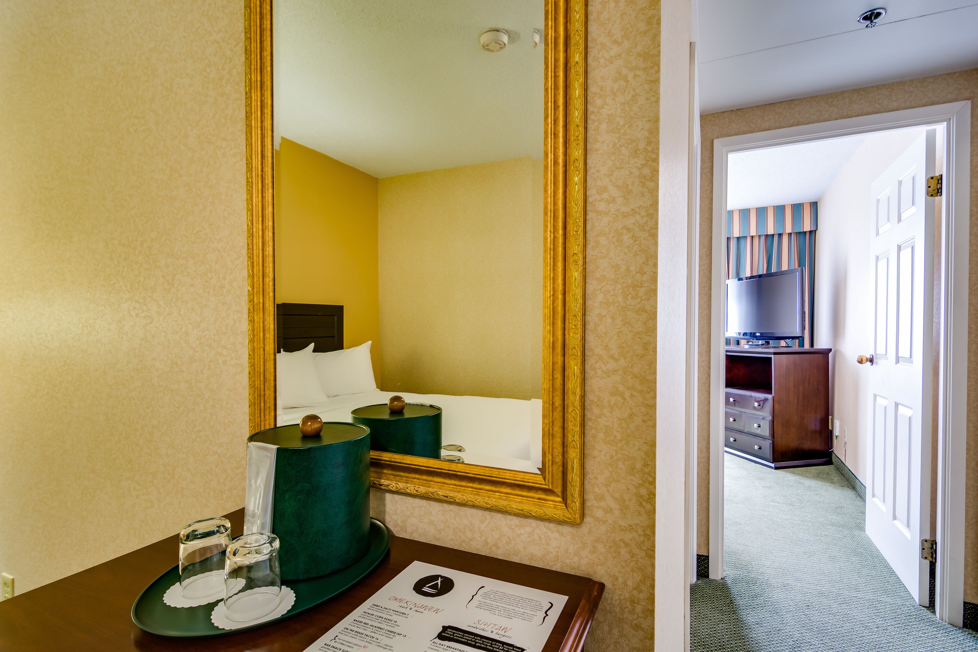 A hotel room with a bed, a mirror, a bedside table with glasses and a tray, and a door leading to a hallway with a refrigerator and bookshelves.
