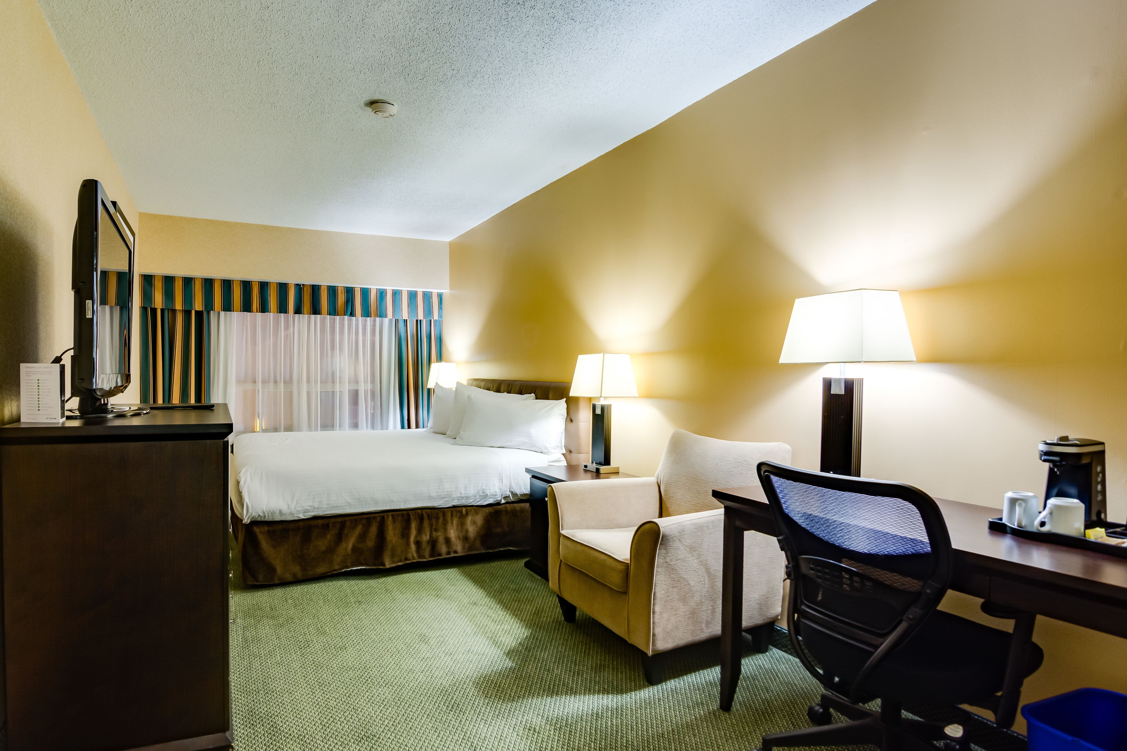 A hotel room with a bed, desk, chair, lamps, and a coffee maker.